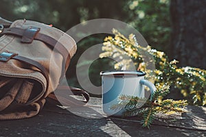 Enameled cup of coffee or tea, backpack of traveller on wooden board in forest outdoors