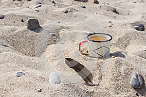 Enameled mug with clean drinking water from Lake Baikal on the shore of the lake and black seagull feather in the sand.