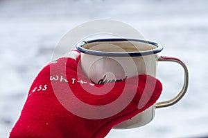 Enameled mug with hot tea in a hand in a red warm mitten in winter on a snowy background.