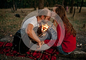 Enamored couple sits on picnic in forest with guitar on background of bonfire flame.