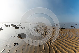 Endless ocean floor uncovered at low tide with sand structures and rocks and tidal pools under lifting fog in the blue sky