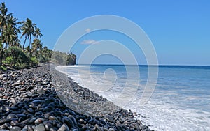 An endless virgin pebble beach with palm trees and tropical vegetation on Bali Island in Indonesia. The waves wash the stony coast