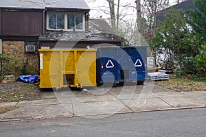 The ends of two dumpsters in the driveway of a house that is having its roof shingles replaced