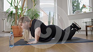 Endurant plus-size Caucasian sportswoman doing push-ups on exercise mat at home. Side view portrait of confident plump