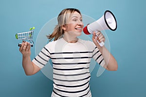 energetic blond attractive young woman in casual outfit announces sale using loudspeaker holding grocery cart on blue