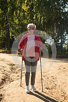 energetic elderly female in red jacket and headphones walking on the beach sunny day using Nordic poles.