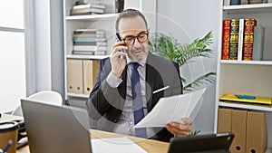 Energetic middle age man with grey hair, immersed in his professional role at the office, confidently talking on a smartphone