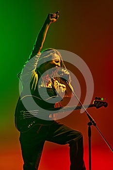 Energetic soulful man with dreadlocks, musician with electric guitar singing against gradient red green background in