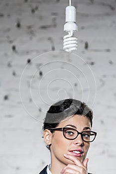 Energy efficient lightbulb hanging over young thoughtful businesswoman against brick wall at office