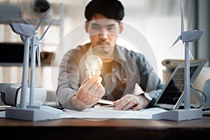 Energy engineer holding light bulb in modern office with solar cell sample and wind turbine model, Creative thinking innovative