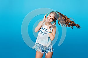 Energy girl with blue headphones  listening to music on blue background in studio. She wears white T-shirt, shorts. Lon