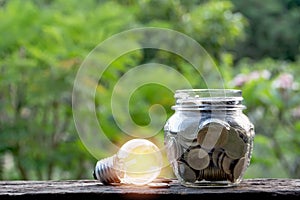 Energy saving light bulb and coins in glass jar on nature background. Saving, accounting and financial concept.