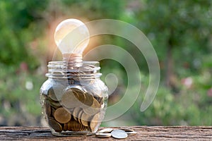 Energy saving light bulb and coins in glass jar on nature background. Saving, accounting and financial concept.