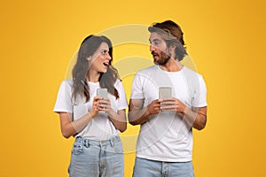 Engaged couple holding smartphones, the woman looking surprised and speaking to the man