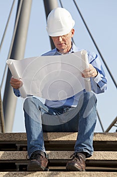 Engineer or architect with hard hat reading a plan while sitting in front of construction of steel