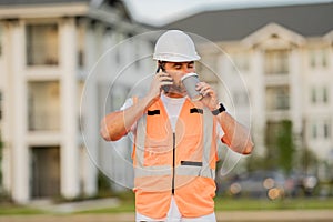 Engineer builder drinking take away coffee using phone on break. Builder at construction site. Buider with helmet on