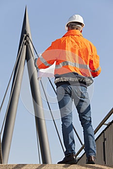 Engineer or foreman with hard hat and protective workwear reading a plan in front of industrial background