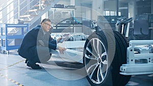 Engineer with Glasses Works on a Tablet Computer Next to an Electric Car Chassis Prototype with Wh