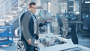 Engineer with Glasses Works on a Tablet Computer Next to an Electric Car Chassis Prototype with Wh