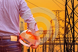 Engineer holding yellow safety helmet with high voltage electric pylon pillars in electric power plants substation on sunset