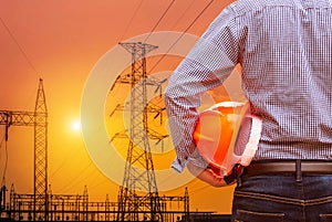 Engineer holding yellow safety helmet with high voltage electric pylon on sunset