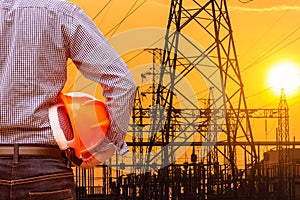 Engineer holding yellow safety helmet with high voltage electric pylon on sunset