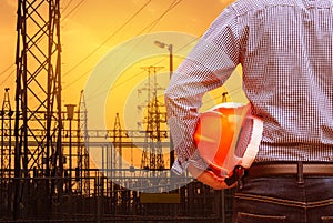 Engineer holding yellow safety helmet with high voltage electric pylon on sunset