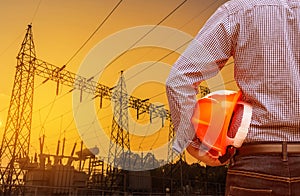 Engineer holding yellow safety helmet with high voltage electric pylon on sunset