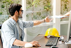 Engineer man receive the document from co-worker and sit in front of computer on the desk in glass window office