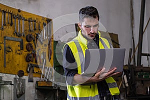 Engineer man wearing safety vest standing in the factory while holding laptop and looking on screen. Worker inspection and