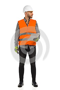 Engineer in orange reflective waistcoat and white helmet is standing with documents and looking at the side