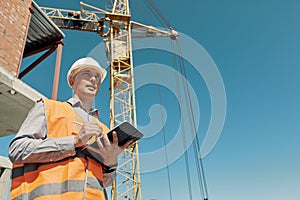 An engineer in an orange vest and a white construction control helmet conducts an inspection with a tablet in his hands
