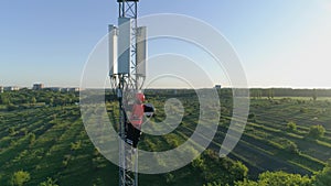 Engineer in safety vest and hard hat climbs high on cellular antenna, drone view of telecommunication tower