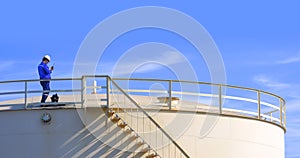 Engineer using walkie talkie while working to check oil quality on top of fuel storage tank against blue sky