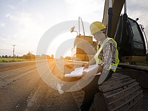 Engineer wear a hard hellmet and holding blueprint on road construction site with machinery