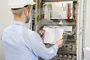 Engineer in white helmet reads design drawing against electric industrial panel. Service worker analyzes the electrical circuit