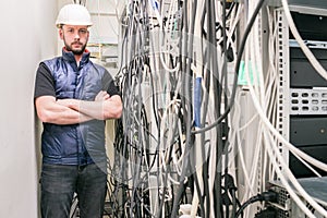 Engineer in a white helmet stands near computer racks with a lot of tangled wires. The man is behind the server cabinets. The