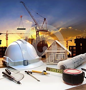 engineer working table plan, home model and writing tool equipment against building construction crane with evening dusky sky