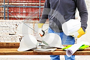 Engineering hand holding white safety hat and paper plan blueprint on wood table