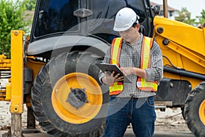 Engineering wearing a white safety helmet standing In front of the backhoe And are using tablet for check the blueprint with const