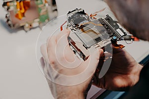 Engineers hands holds broken photo camera, poses against workspace, repairs digital device or professional dslr camera.