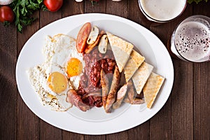 English breakfast (fried eggs, beans, roasted bacon, sausages and vegetables) on dark wood background