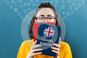 English language day. Portrait of a young woman holding english dictionary, covering half of her face. Blue background with