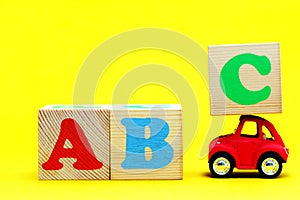 English letters ABC on wooden blocks on a yellow background. A toy car carrying a block with the letter C on the roof