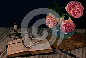 English rose in a vase on a table next to an open book with glasses for reading and a burning candle with a dark background