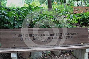 An engraved wood park bench with a quote from Robert Herrick in front of a shade garden with trees and plants