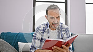 Engrossed middle age man with grey hair, comfortably sitting on his living room sofa, absorbed in reading a book at home