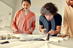 Enhance the quality of dressmaking. Young male designer cutting white fabric textile in a studio. Group of creative