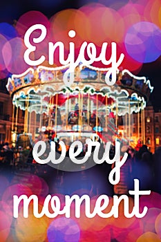 ENJOY EVERY MOMENT. Inspirational Typographic Quote. Merry-Go-Round illuminated at night with colorful bokeh.