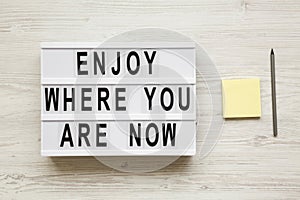 `Enjoy where you are now` words on lightbox over white wooden background, overhead. Top view, flat lay, from above